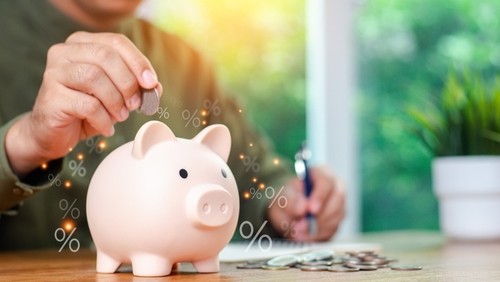 A person placing a coin into a pink piggy bank on a wooden table, with floating percentage symbols representing savings or interest rates. In the background, a notebook, pen, and a potted plant are visible near a window with natural light.